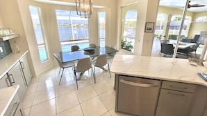 A modern kitchen with a glass dining table, a built-in dishwasher, and large windows offering a view of a body of water. - TOP FLORIDA's Villa Lake View (Cape Coral)