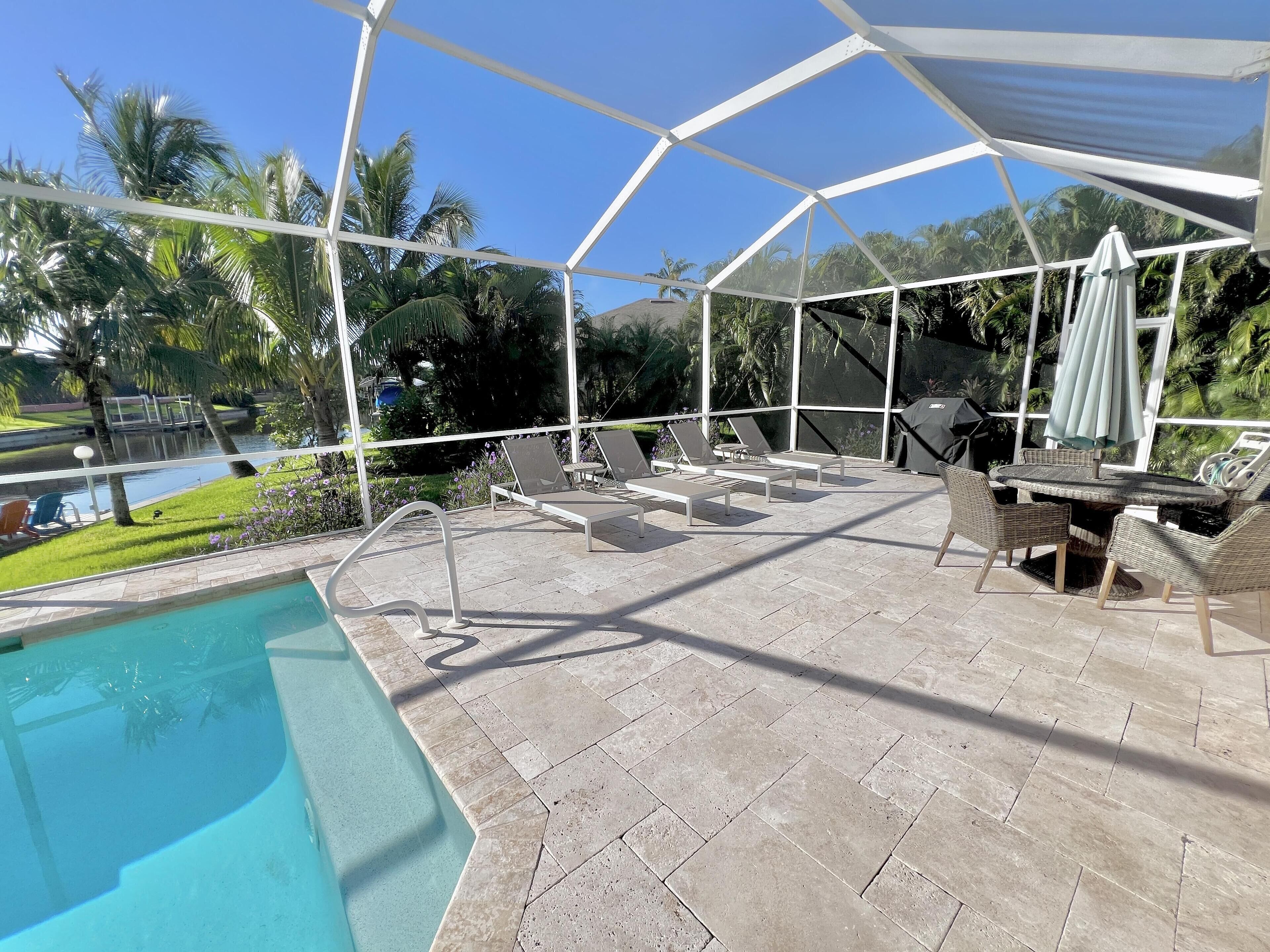 An outdoor pool area with lounge chairs and a table, surrounded by a covered patio with a view of the pool and greenery.
