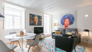 A modern living room with a round dining table, grey chairs, a grey sofa with yellow cushions, a colorful rug, and two large paintings on the walls. - FLH Chiado Modern Place (Lisbon)
