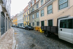 A cobblestone street lined with colorful buildings, parked cars, and a white truck.