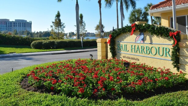 Schild für 'Sail Harbour' mit Blick auf den Strand, Palmen und einem Gebäude im Hintergrund.