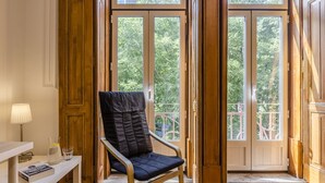 Ein Zimmer mit Holzboden, einem schwarzen Sitzsessel, einem weißen Tisch mit Lampe und Blick durch große Glastüren auf einen Außenbalkon.