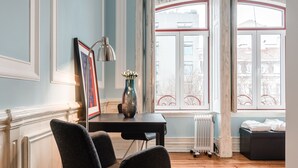 Una habitación con piso de madera, una mesa negra con una lámpara, dos sillas, un florero con flores y una ventana con vista a edificios.