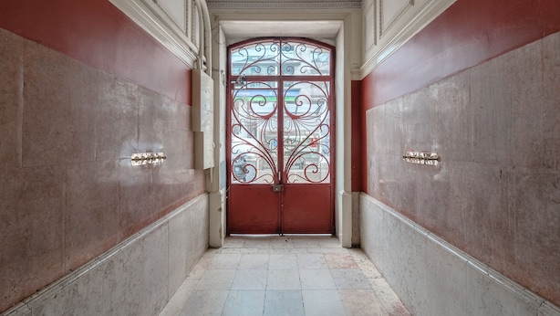 Un pasillo con una puerta roja, adornada con detalles de metalurgia, que da a una habitación con techo blanco y paredes de mármol.