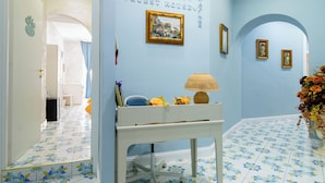 A hallway with blue walls and floral patterned floor tiles, a white console table with a lamp, and framed pictures on the wall. - Alma Adry (Naples)