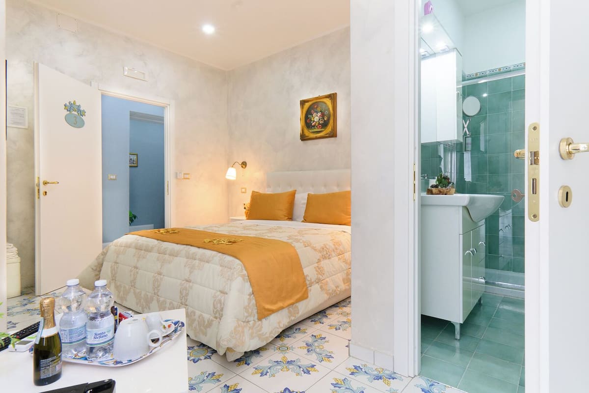 A hotel room with a bed, a bathroom with a glass shower, and a small table with bottles and a remote control.