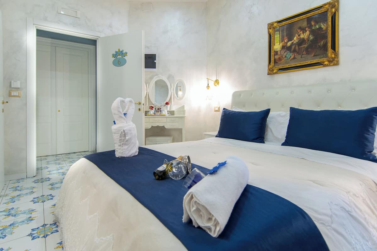 A hotel room with a large bed, a white headboard, a painting on the wall, a vanity with a mirror, and a door with a number sign.