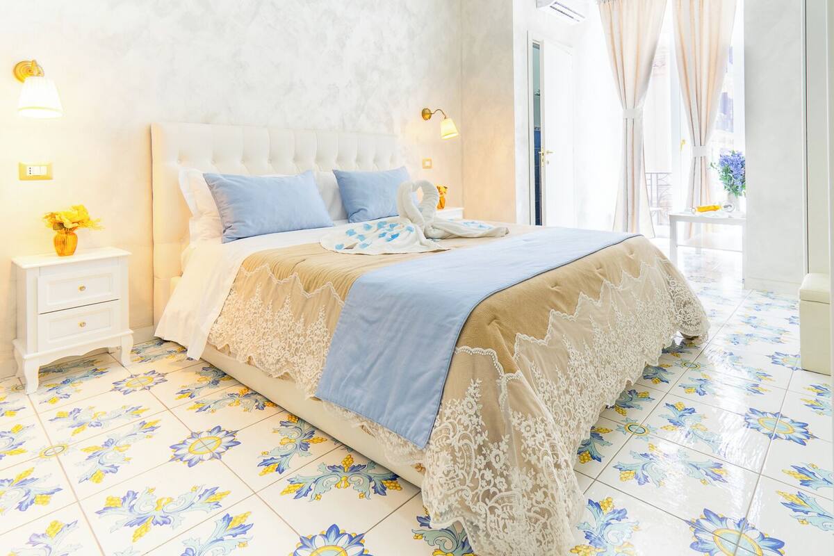 A bedroom with a tufted headboard, a floral-patterned floor, a bedside table with a lamp, and a window with curtains.