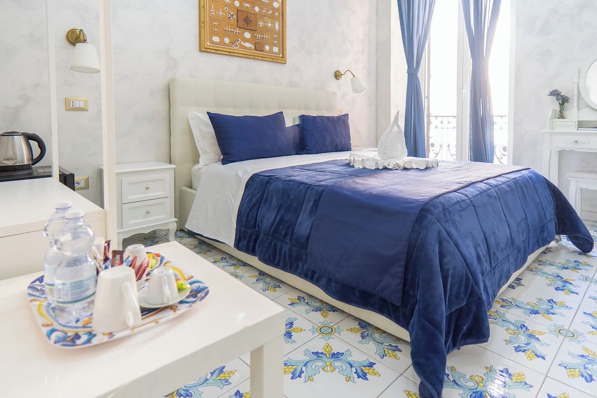 A hotel room with a large bed, a bedside table with a tray of toiletries, a white nightstand, and a wall-mounted light fixture.