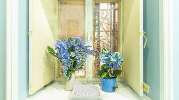 A narrow entrance with a white door and a metal gate, leading to a small porch area with a white bench and two potted plants with blue flowers.