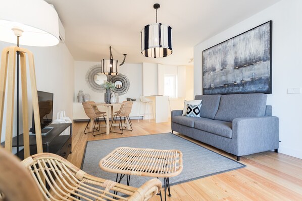 A modern living room with a grey sofa, wicker chairs, a dining table with chairs, a large abstract painting, and a hanging light fixture.