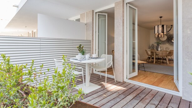 Une terrasse extérieure moderne avec une table et des chaises blanches, une terrasse en bois et une vue sur l’espace repas intérieur.