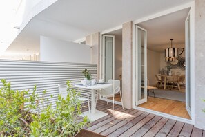 A modern outdoor patio with a white table and chairs, a wooden deck, and a view of the interior dining area.