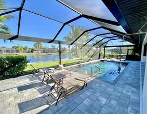 A covered outdoor swimming pool with lounge chairs and a view of a water body and palm trees.