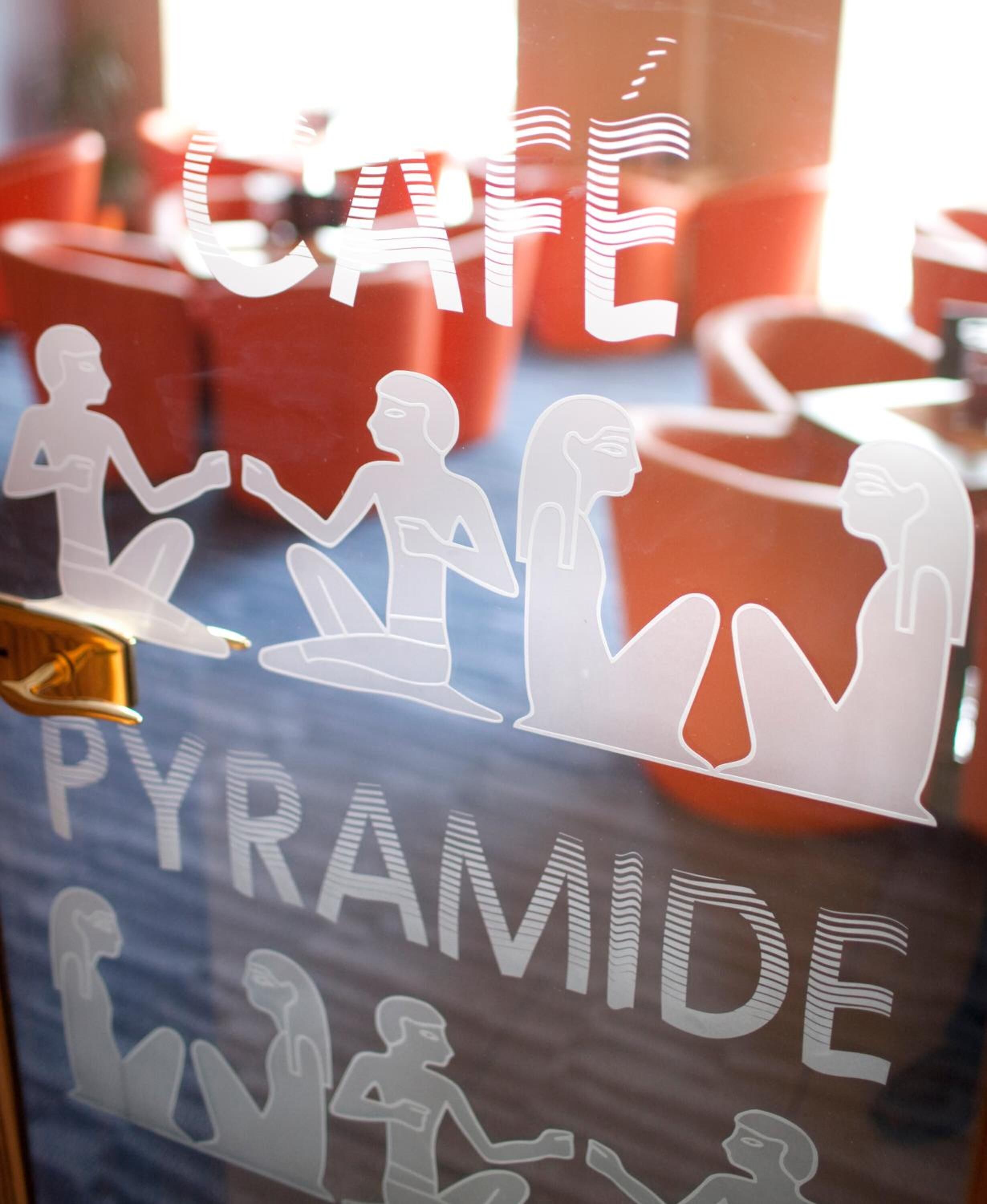 Una ventana con la inscripción "CAFE PYRAMIDE" y siluetas de personas en diferentes poses.