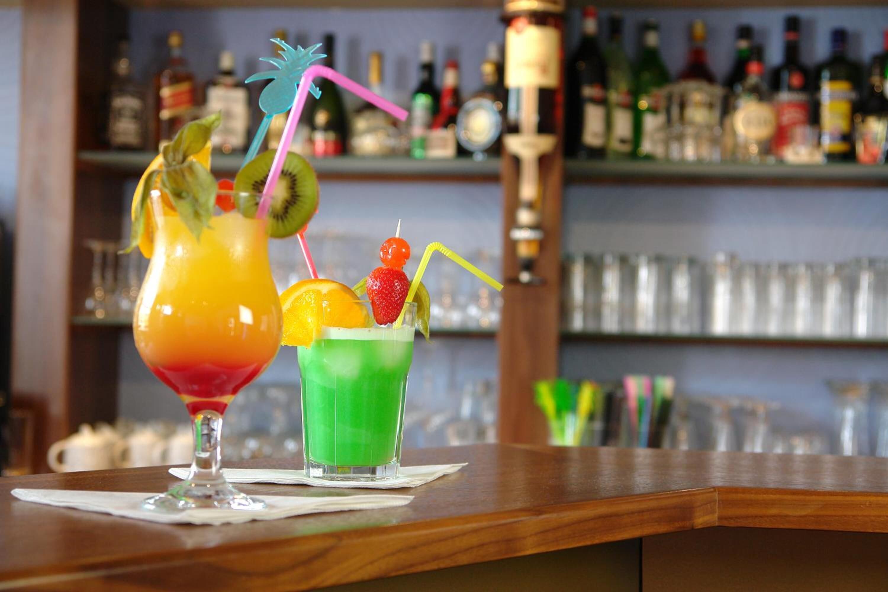 Zwei Cocktails auf einer Bar, im Hintergrund eine Flaschenregal mit Spirituosen.