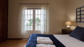 Ein Schlafzimmer mit Bett, Nachttisch, Lampe und Blick durch ein fenster mit Vorhängen.