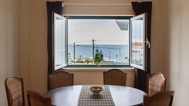 Ein Essbereich mit einem runden Tisch, sechs Stühlen, einem Fenster mit Vorhängen und Blick auf das Meer und Gebäude.
