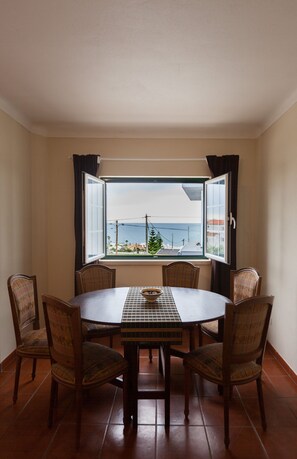 Un comedor con una mesa redonda y seis sillas, una ventana con cortinas, y vistas al mar y edificios.