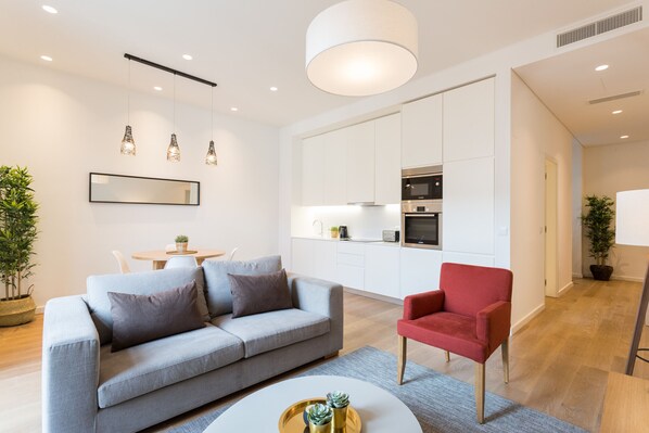 A modern living room with a grey sofa, a red armchair, and a round coffee table. There is a kitchen area with white cabinets and a dining table with chairs.