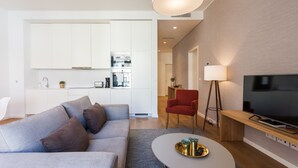 A modern living room with a grey sofa, a round coffee table with decorative items, a red armchair, a TV on a wooden stand, and a kitchen area with white cabinets and a large pendant light.