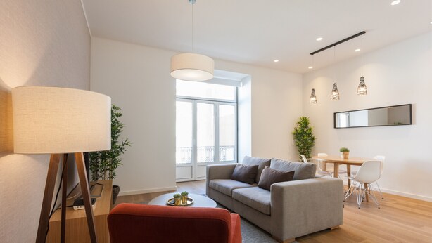A modern living room with a sofa, a red chair, a dining table with chairs, a lamp, and a large window.
