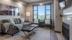 A modern living room with a sofa, ottoman, and chairs, a fireplace, a TV mounted on the wall, and large windows offering a view of the outdoors.