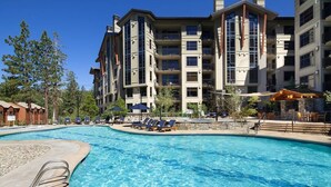 A swimming pool surrounded by a multi-story building with balconies, lounge chairs, and a clear sky.