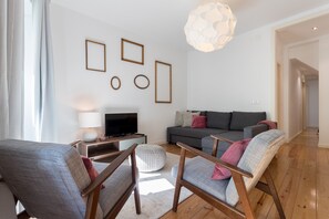 A modern living room with a grey sofa, wooden chairs, a TV, and wall art.