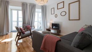 Un soggiorno moderno con un divano grigio, una sedia in legno, un mobile TV e un'ampia finestra con tende.