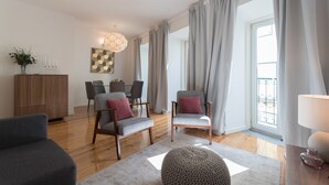 Un soggiorno moderno con pavimento in legno, divani grigi, un poggiapiedi rotondo e una zona pranzo con tavolo e sedie.