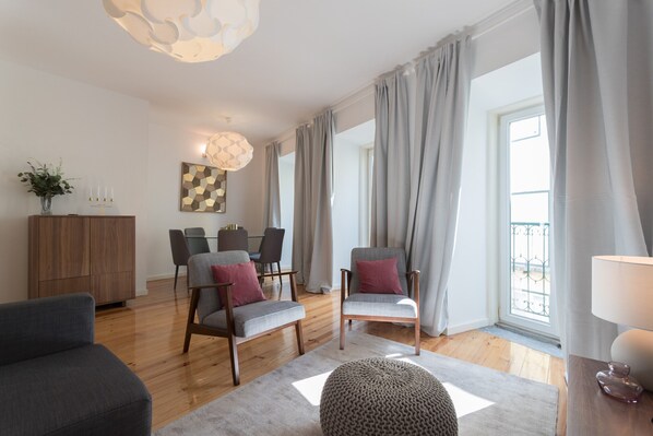 Una sala de estar moderna con piso de madera, sofás grises, un otoman redondo y un comedor con mesa y sillas.