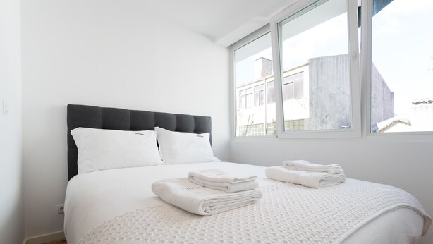 Un letto rifatto con lenzuola bianche e una testata grigia, una finestra con vista sugli edifici e un tappeto bianco con trama sul pavimento.