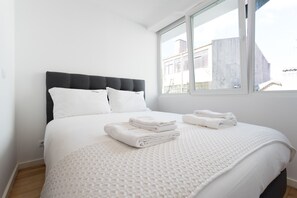 A neatly made bed with white linens and a grey headboard, a window with a view of buildings, and a white textured rug on the floor.