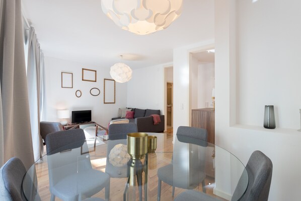 A modern living room with a glass dining table, grey sofas, and a TV.