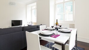 Uma área de refeições com uma mesa branca posta para dois, um sofá cinza e uma televisão ao fundo.
