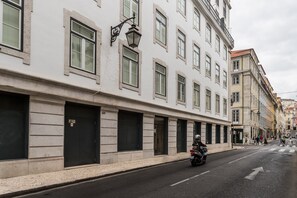 Une rue bordée de bâtiments blancs, avec des devantures de magasins fermées, et un motocycliste qui roule sur la chaussée.