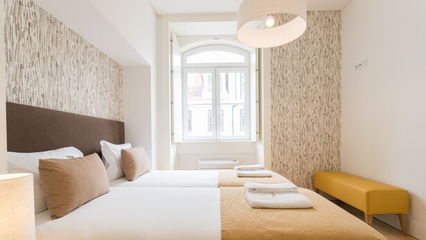 A hotel room with two beds, a bench, a lamp, and a view of a building through a window. - FLH Downtown Fantastic Flat (Lisbon)