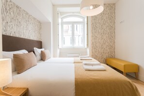 A hotel room with two beds, a bench, a lamp, and a view of a building through a window.