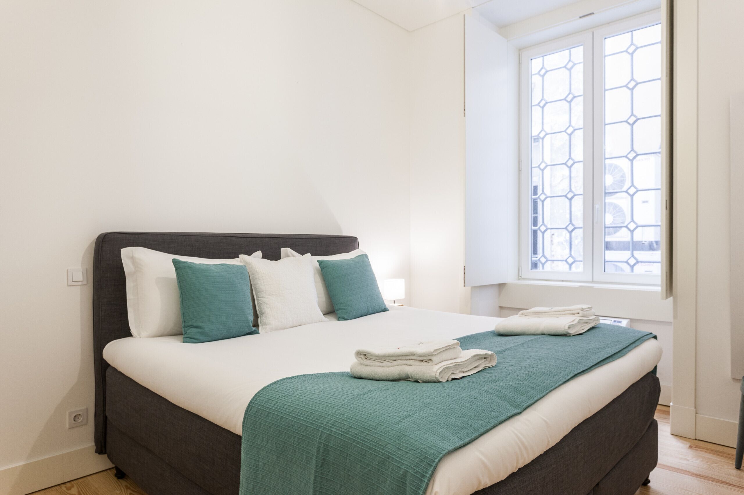 Una cama bien hecha con ropa de cama blanca y aguamarina, un cabecero oscuro y una ventana con un diseño geométrico.