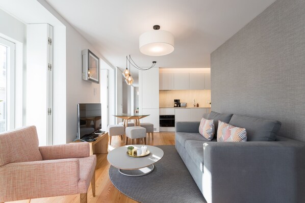 Un salon moderne avec un canapé gris, un fauteuil rose, une table basse ronde et un coin repas comprenant une table et des chaises en verre.