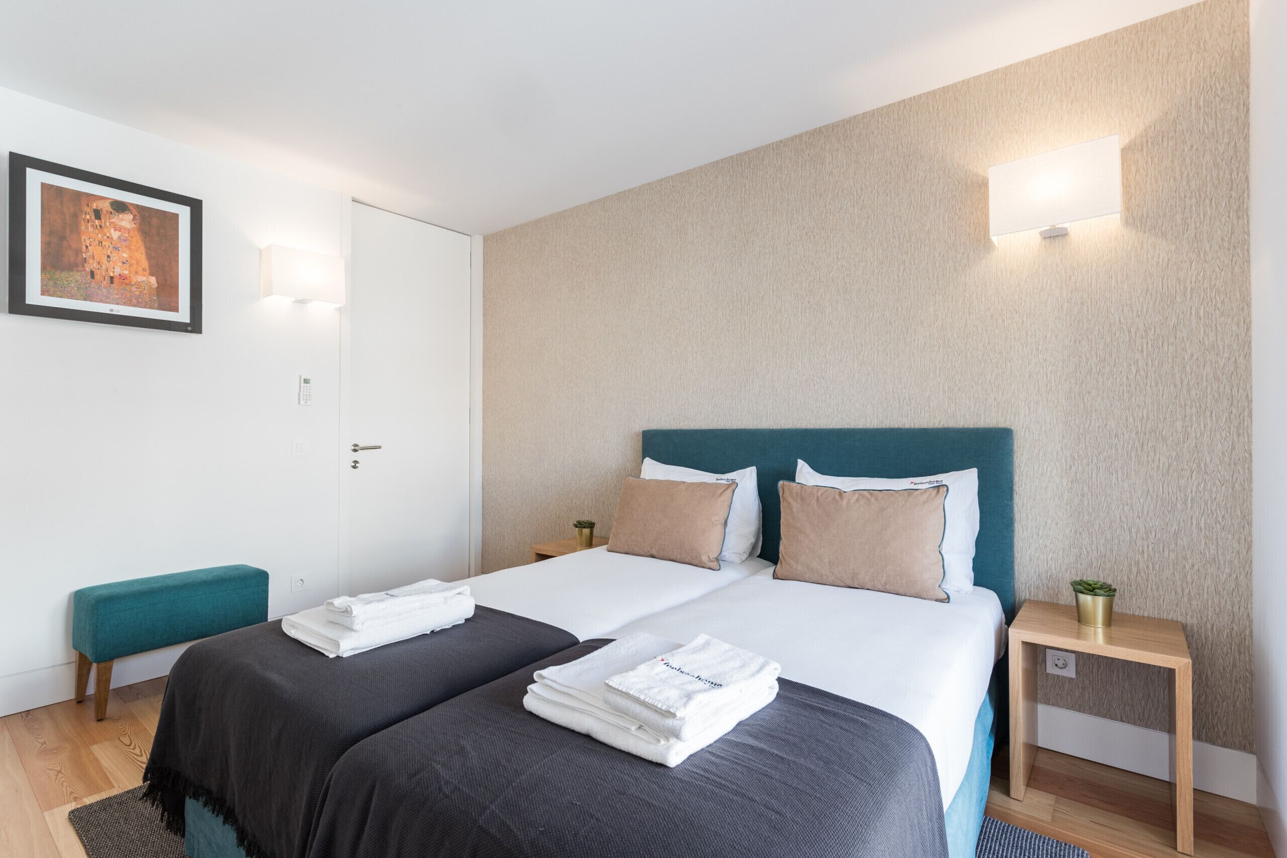 A hotel room with two beds, a bench, a bedside table with a plant, and a painting on the wall.