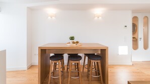 A modern kitchen with a wooden table and three bar stools, a small plant on the table, and recessed lighting. - FLH Chiado Bright Apartment (Lisboa ciudad)