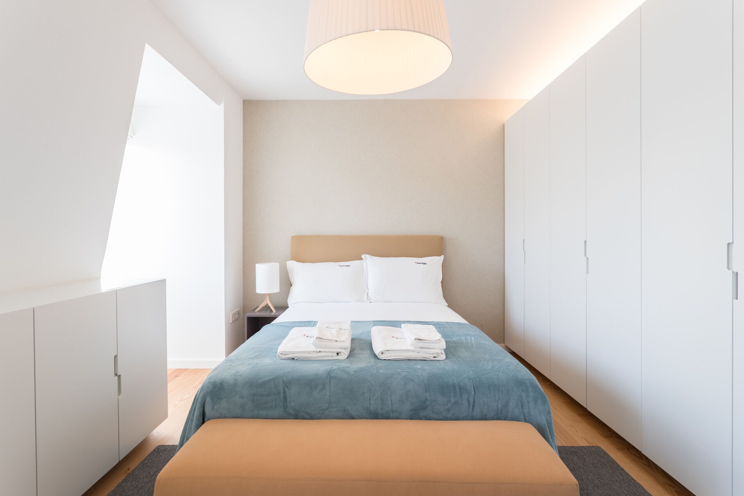 A modern hotel room with a large bed, a bedside table with a lamp, and a wardrobe.