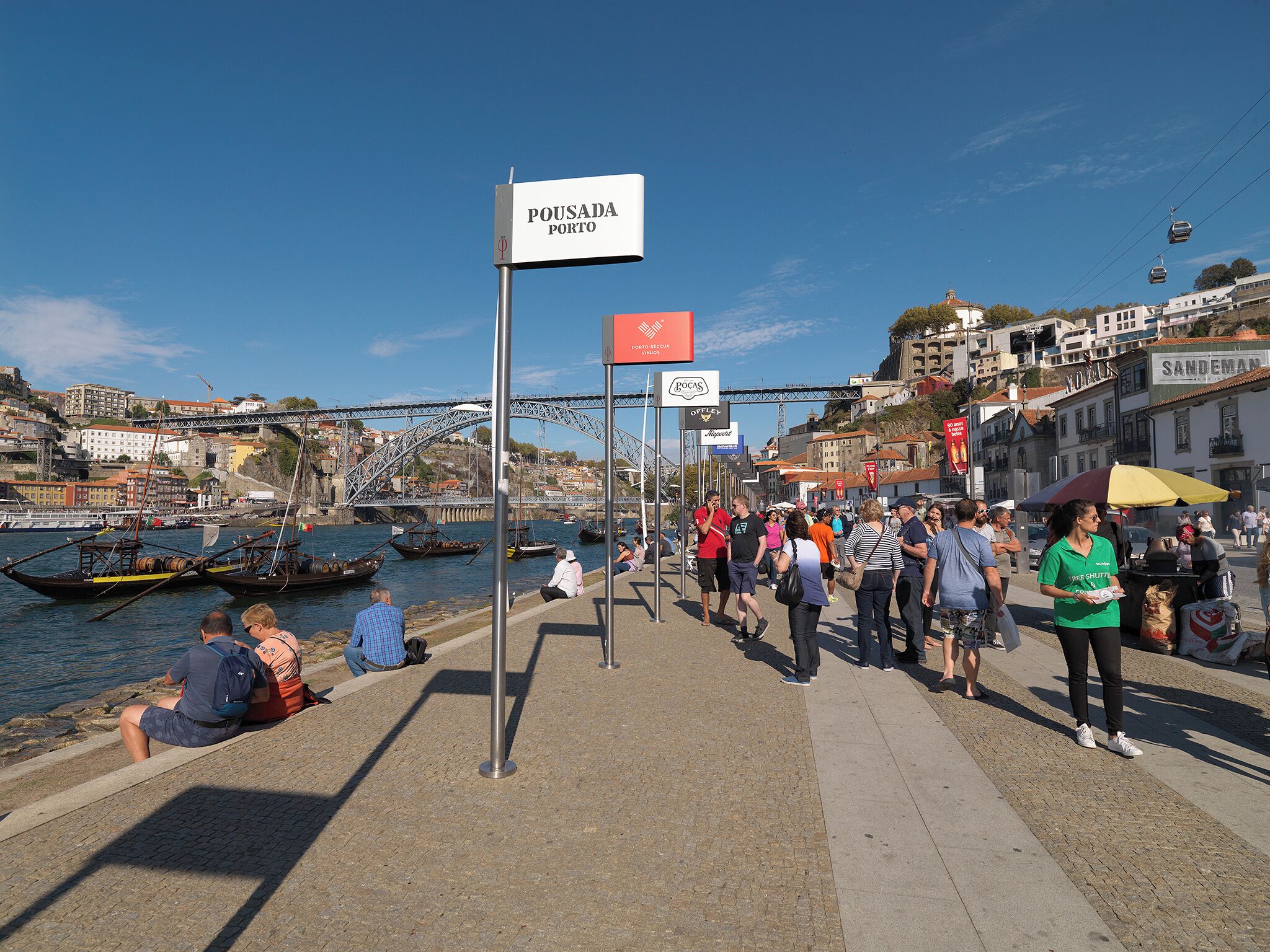 A waterfront promenade with a sign indicating 'Pousada Porto', a bridge in the background, and boats docked along the river.