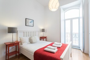 A bedroom with a bed, two bedside tables, a red blanket, a framed picture, and a balcony with a railing.