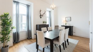 Een eetkamer met een donker houten tafel, zes stoelen, een kroonluchter, een decoratie van een hertenkop, een potplant en een ingelijst beeld aan de muur.