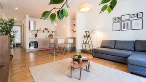 Una sala de estar moderna con un sofá gris, una mesa de centro de madera con una planta, y una cocina al fondo.