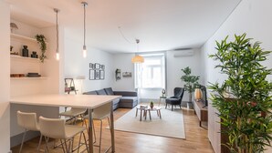 Una sala de estar moderna con sofá, mesa de centro y comedor. Hay plantas, una lámpara y una ventana que proporciona luz natural.