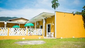 A yellow building with a covered patio area, white railings, and a palm tree. - Recanto Soga Chalés (Ubatuba)